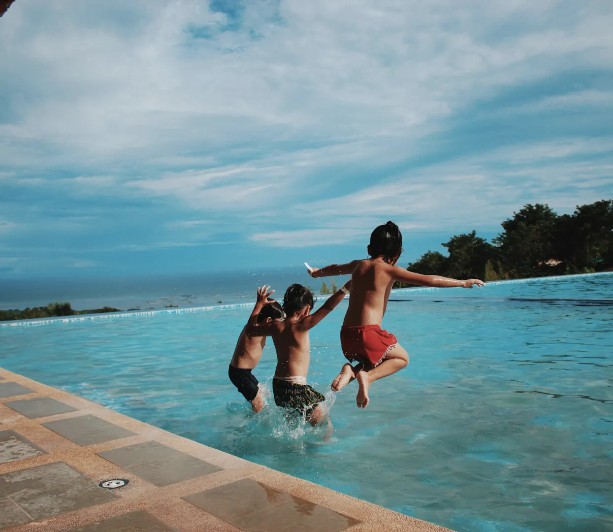 Three kids jumping all at once into an infinity pool at a beachside hotel.